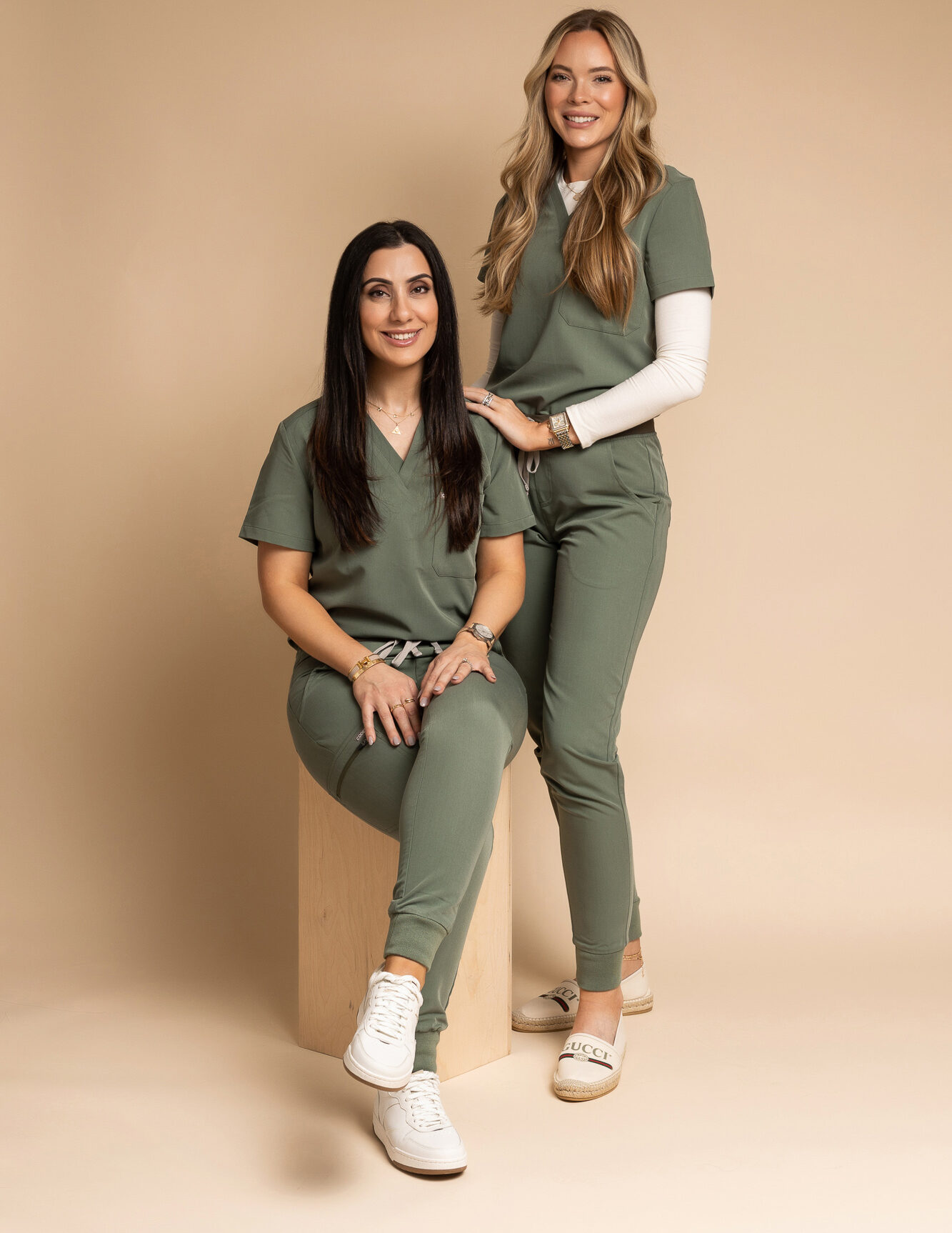 One women sits while another stands near her. Both smile into the camera.