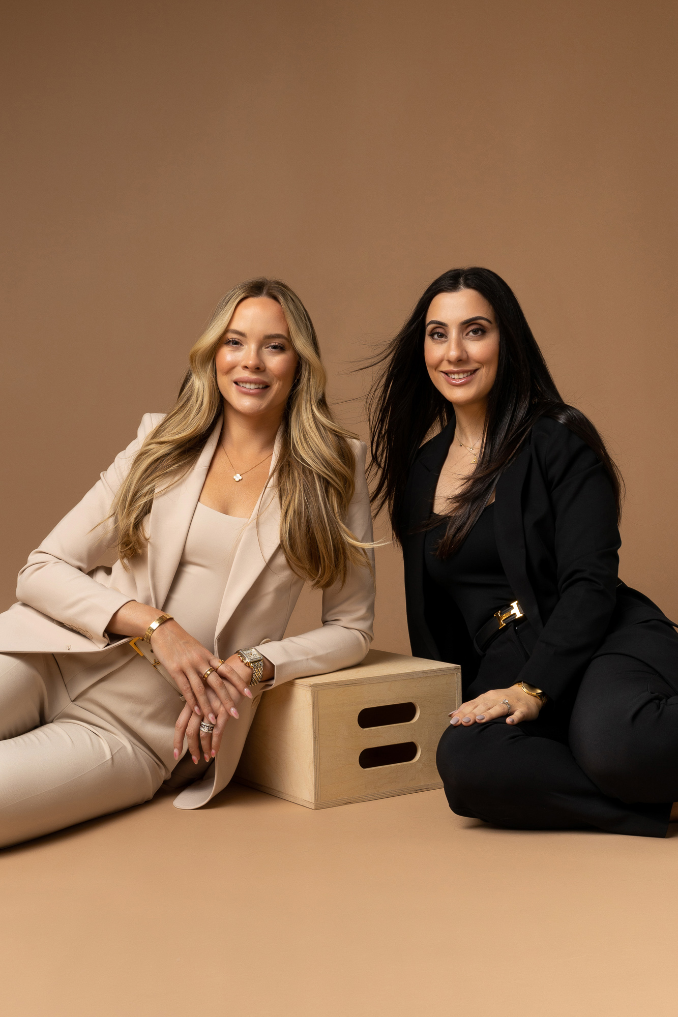 Two women sit on the ground while smiling into the camera.