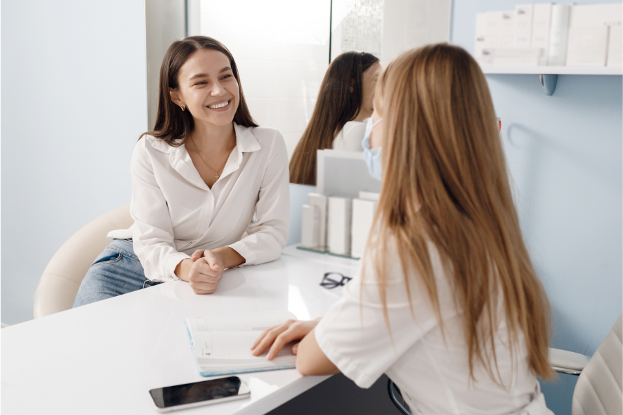 A young brunette patient talking to an injector during her consultation at Exhibit Medical Aesthetics, smiling confidently after learning how she can benefit from receiving fillers near Madison, NJ.