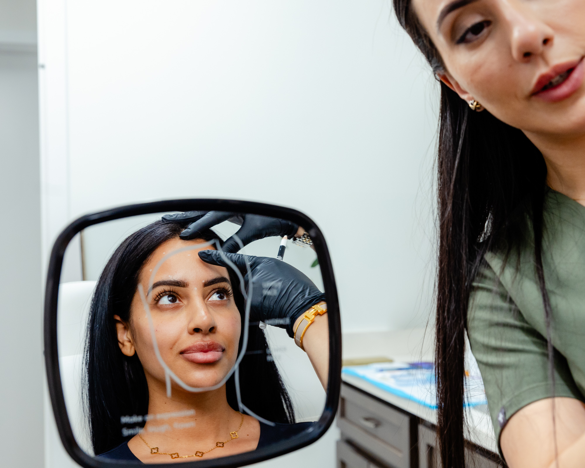 A young dark-haired female patient at Exhibit Medical Aesthetics holding a mirror in front of her face while her female injector shows her exactly where she will place the injections of Botox near Madison, NJ.