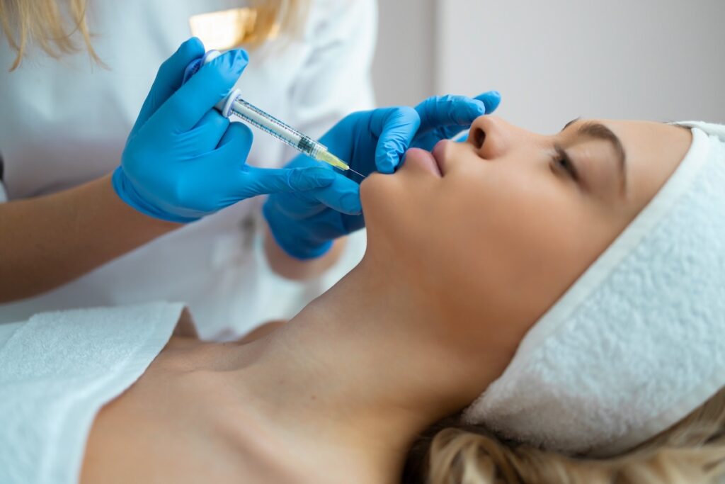 Side angle of woman lying down as nurse injects filler needle into her chin.