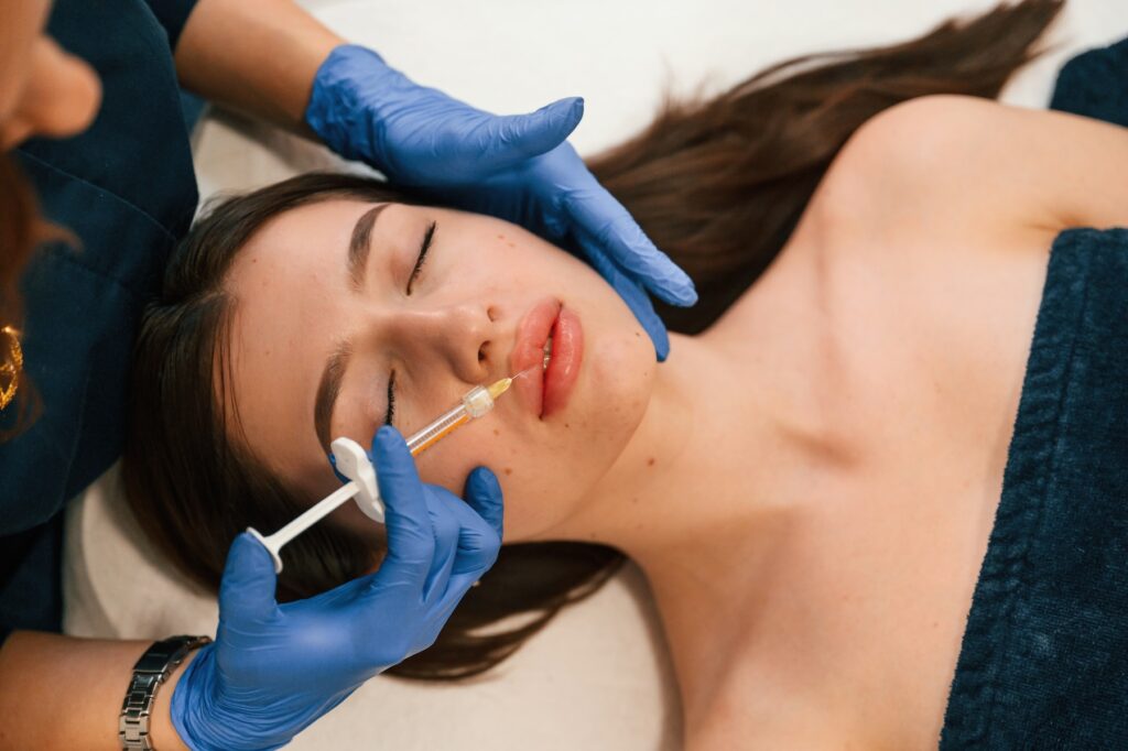Woman lying down with eyes closed as cosmetologist wearing gloves inserts needle into her top lip.