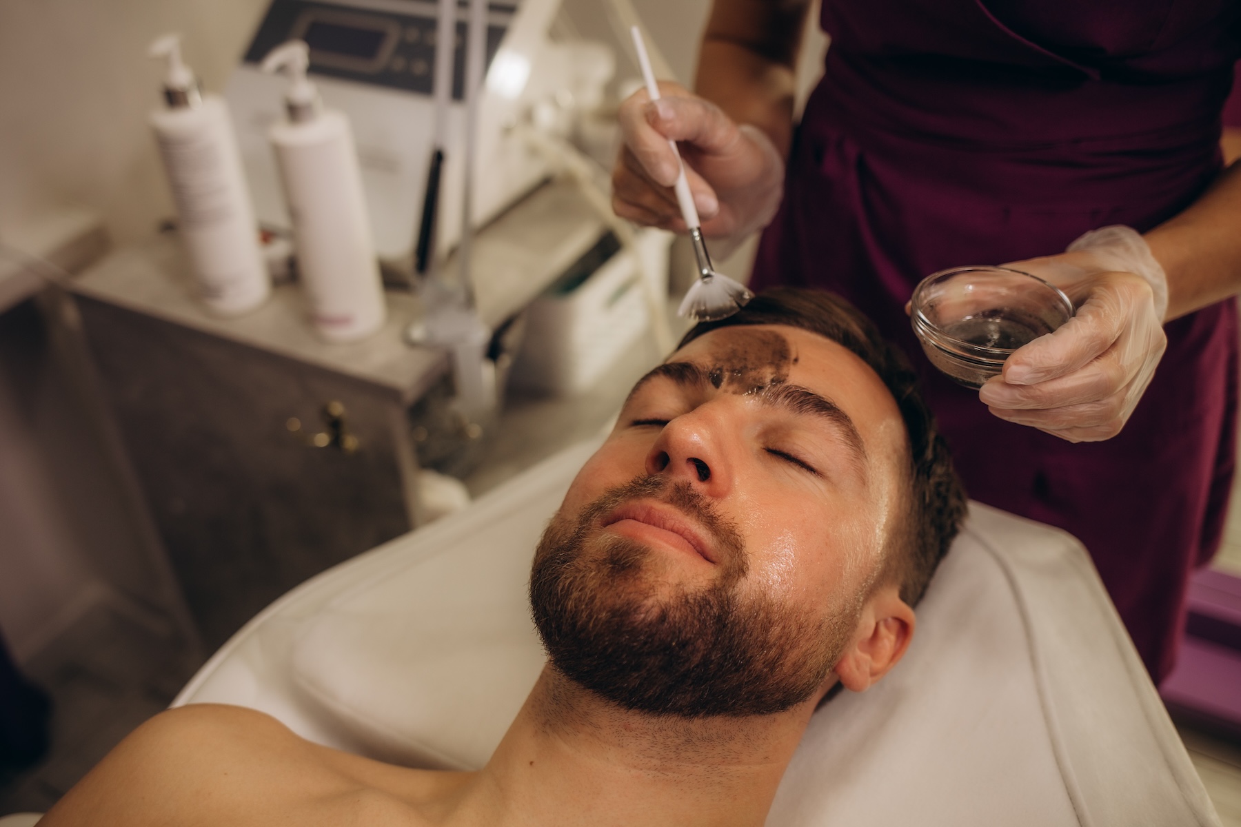 Above angle of relaxed and serene young man keeping head on rolled soft towel while beautician applying clay mask on his face in spa salon. High quality photo