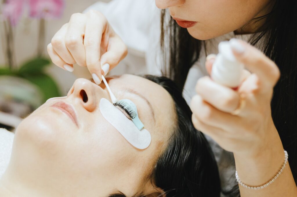 Portrait of a young semi-recognizable Caucasian girl cosmetologist in a white coat removing brown dye from the eyelashes of her left eye using a cotton swab to a brunette woman lying on the cosmetology table in a home beauty salon for eyelash lift, side view close-up with selective focus.