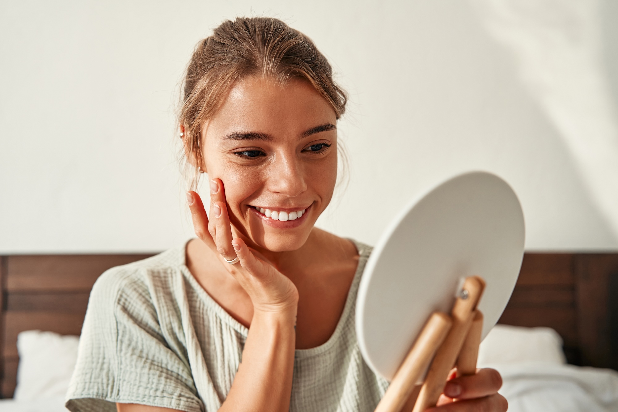 A young blonde woman sitting in bed at home looking happily at her reflections after visiting Exhibit Medical Aesthetics for rosacea treatment in Denville.