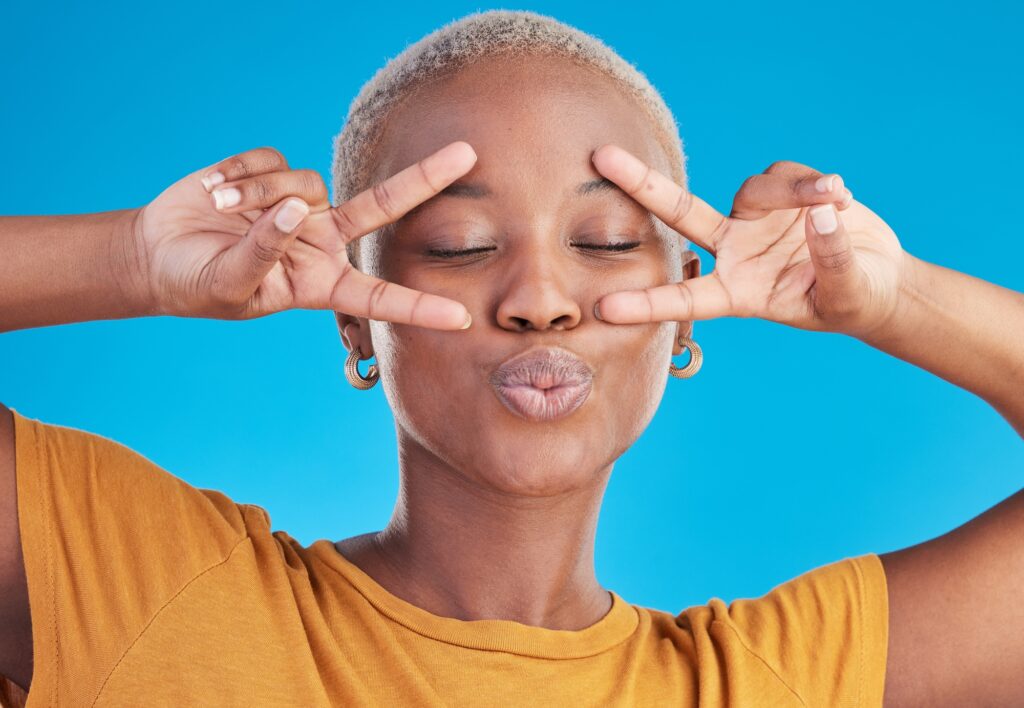 An African-American woman with short blonde hair making a kissy face while holding up peace signs after visiting Exhibit Medical Aesthetics to improve her lip shape using lip filler near Mountain Lakes, NJ,